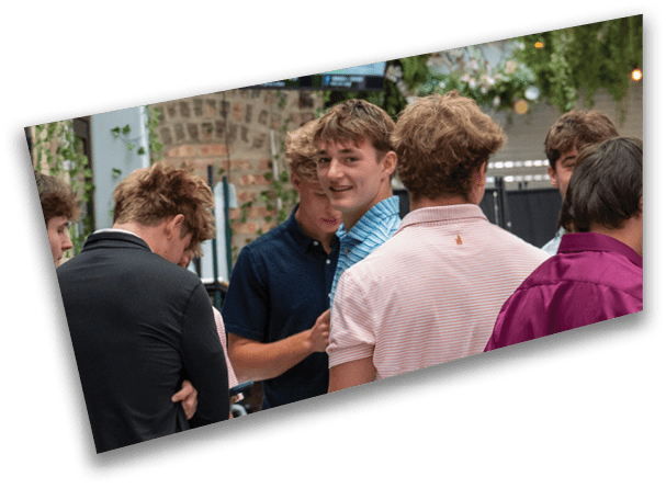 A group of people wearing casual shirts are gathered and engaged in conversation in an outdoor setting with greenery and brick walls. Description generated by AI