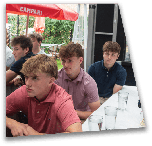 A group of people wearing casual polo shirts are seated closely together at a table with glasses of water in an outdoor setting under a red umbrella. Description generated by AI