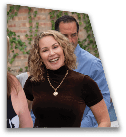 Two adults are standing close together outdoors with a brick wall and green vines in the background, one smiling broadly and the other looking downward. Description generated by AI