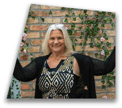 A smiling adult with shoulder-length light hair wearing a patterned dress and black cardigan poses with one arm raised in front of a brick wall decorated with green vines and pink flowers. Description generated by AI