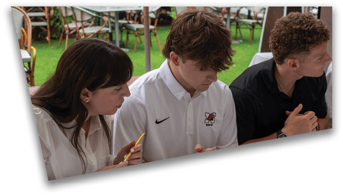 Three people sit closely together at a table, with one person holding a pencil and another wearing a white polo shirt with a logo and a Nike swoosh. Description generated by AI