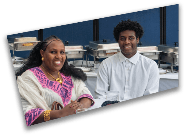 Two people are sitting at a table with food warmers in the background, both smiling, with one wearing a white shirt and the other wearing a white and pink traditional garment with gold jewelry. Description generated by AI