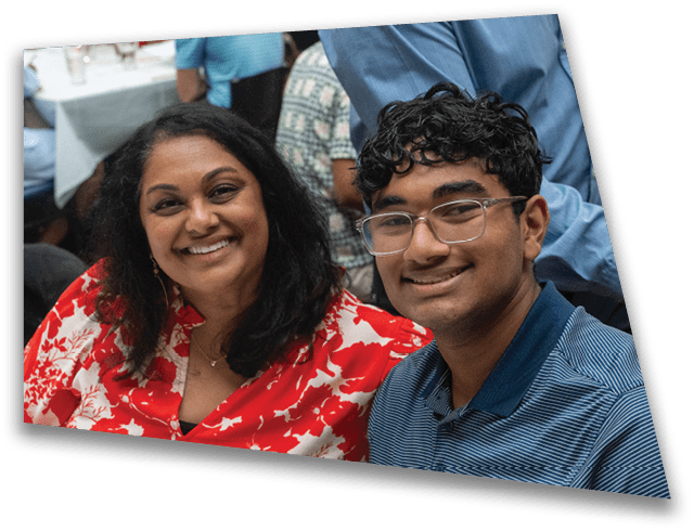 A person with curly hair and glasses is smiling while wearing a blue striped shirt, sitting next to another person in a red and white patterned garment. Description generated by AI