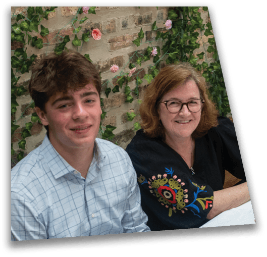 Two people sit closely together smiling in front of a stone wall decorated with green ivy and pink flowers. Description generated by AI