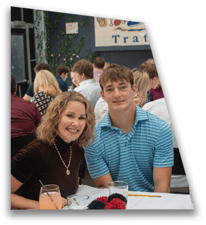 A group of people sitting at tables in a restaurant setting with one person in a blue striped shirt smiling toward the camera. Description generated by AI