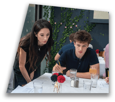 A person with long dark hair wearing a black shirt is leaning over a table while two other people are seated nearby, engaged in a discussion or collaborative activity in a room with a window and green leafy wall decor. Description generated by AI