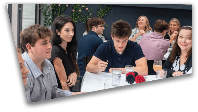 A group of people sitting closely together at a table in a casual dining setting, engaged in conversation and writing. Description generated by AI