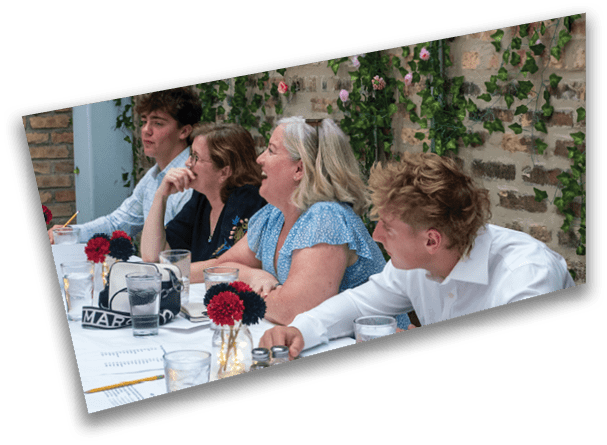 Three adults sit at a table engaged in conversation, with one person wearing a blue patterned dress and another in a white shirt, and a small vase with red and black flowers is on the table. Description generated by AI