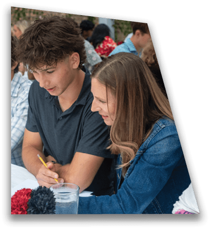 Two people with casual clothing are sitting closely together, one writing with a pencil while the other looks on attentively. Description generated by AI
