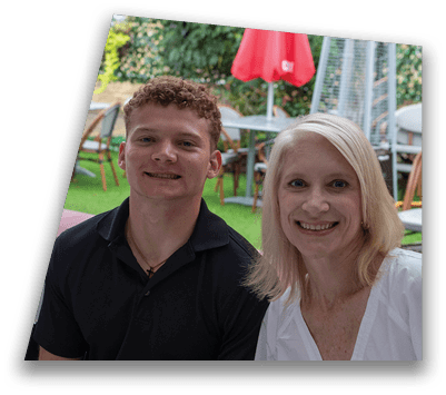 A person wearing a black collared shirt and a gold necklace with a cross pendant is smiling with another person partially visible beside them in an outdoor setting with green grass and wooden chairs in the background. Description generated by AI