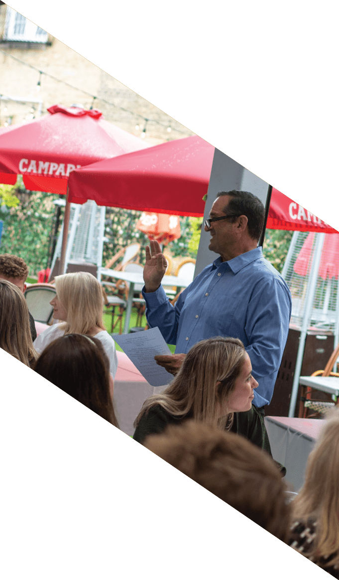 People are seated around a table outdoors under a red umbrella, engaged in conversation and holding papers. Description generated by AI