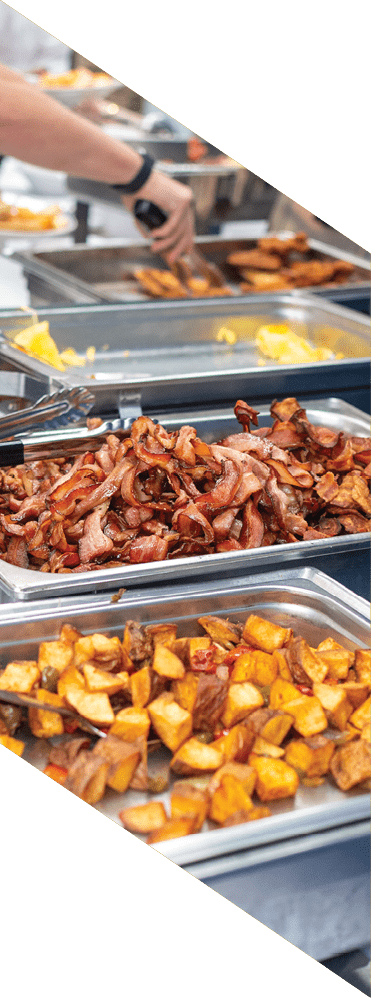 Several trays of cooked breakfast foods including bacon, scrambled eggs, and roasted potatoes are arranged on a buffet table. Description generated by AI