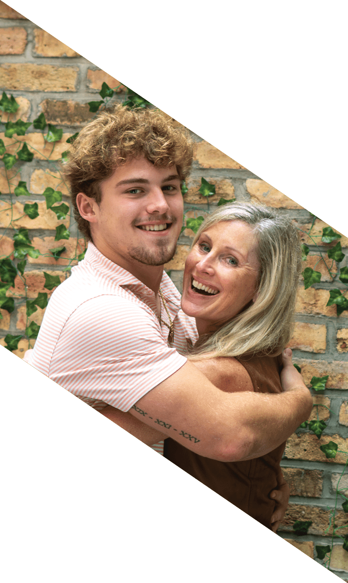 A person with curly hair wearing a light striped shirt is smiling and hugging another person with long hair who is also smiling, with a brick wall and green ivy in the background. Description generated by AI