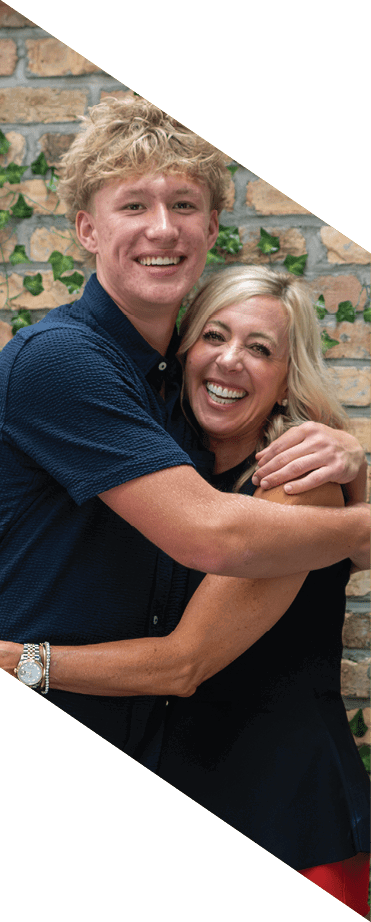Two adults smiling and hugging each other in front of a brick wall decorated with green ivy and pink flowers. Description generated by AI
