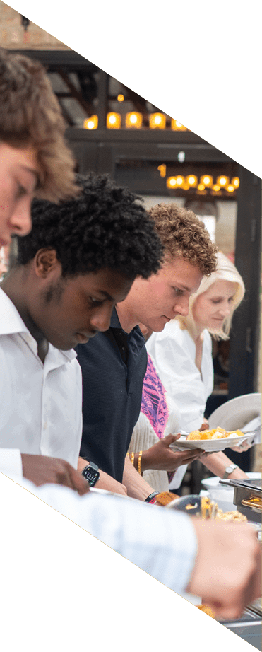Three adults stand in line holding plates while serving themselves food from a buffet in an outdoor setting with greenery and string lights. Description generated by AI