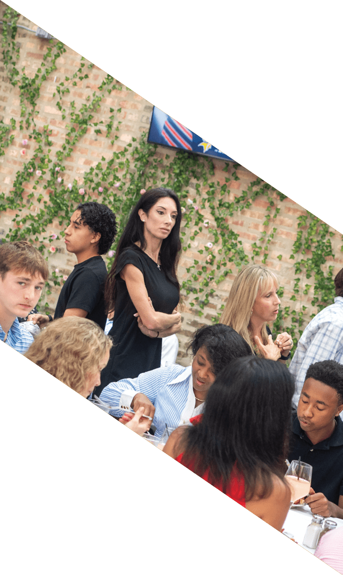 A group of people are seated around a table outdoors, engaged in conversation and dining, with some standing nearby against a backdrop of a brick wall covered in green vines. Description generated by AI