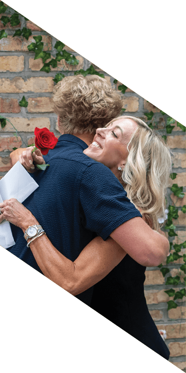 Two people embrace near a brick wall decorated with green vines and pink flowers, with one person holding white envelopes. Description generated by AI