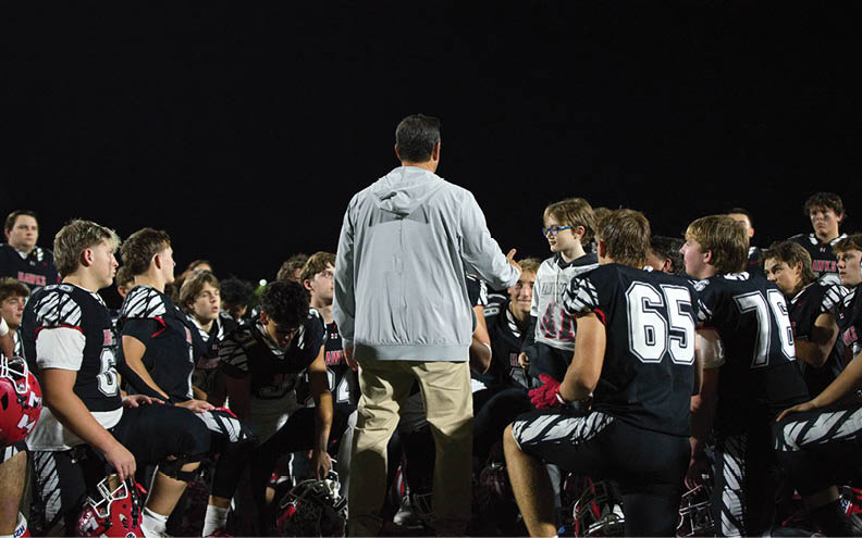 A group of people wearing football uniforms sit on benches outdoors at night while a person in a gray jacket stands in front of them speaking. Description generated by AI