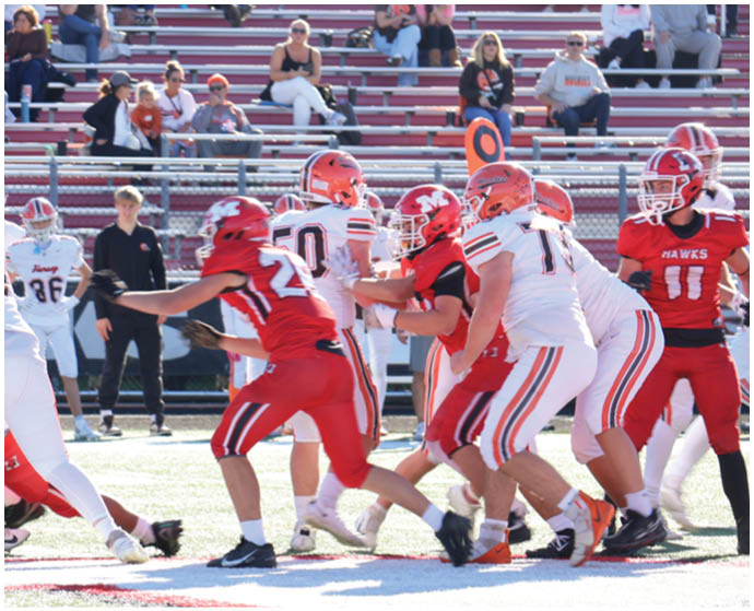 A group of people in football uniforms are actively engaged in a play on a field with spectators watching from bleachers in the background. Description generated by AI