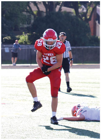 A person in a red football uniform is standing on one leg while another person in a white football uniform lies on the ground on a sunlit field with a referee and spectators in the background. Description generated by AI