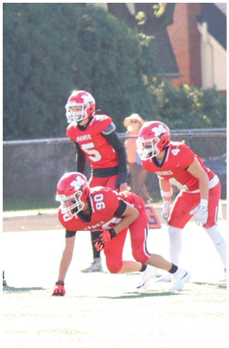 Three people in football uniforms are positioned on a field, with two wearing red jerseys and helmets labeled “HAWKS" and one in a white jersey and helmet crouched in front of them. Description generated by AI