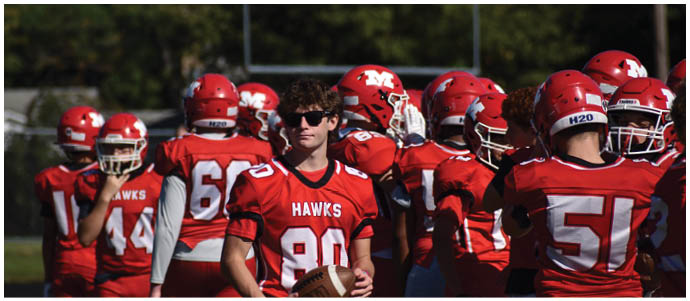A group of people wearing red football uniforms with “HAWKS" on the jerseys are gathered on a field, with one person holding a football and wearing sunglasses. Description generated by AI