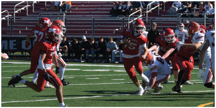 Players in red and white football uniforms are actively engaged in a play on a football field with spectators seated in the background. Description generated by AI