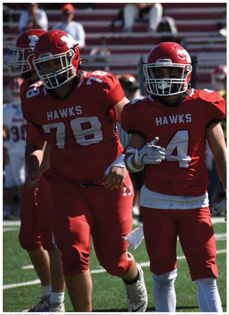 Two people wearing red football uniforms with helmets stand on a football field with blurred spectators in the background. Description generated by AI