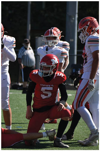 A group of people in football uniforms are on a field with one person in a red uniform kneeling while others in white uniforms stand nearby. Description generated by AI