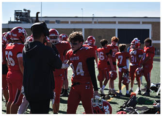 A person in a black jacket is speaking into a headset microphone while a person in a red football uniform with the number 14 and the word “HAWKS" stands nearby on a sports field with other players in red uniforms in the background. Description generated by AI