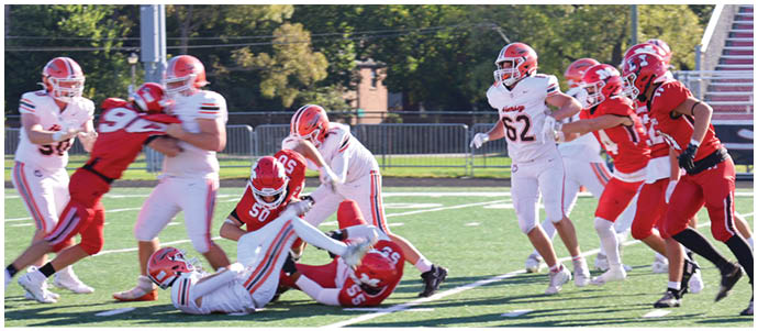 People wearing red and white football uniforms are actively engaged in a play on a grassy field with bleachers and trees in the background. Description generated by AI