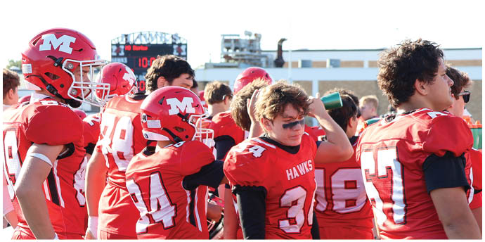 Several people wearing red football uniforms and helmets stand together on a field with a scoreboard and buildings in the background. Description generated by AI
