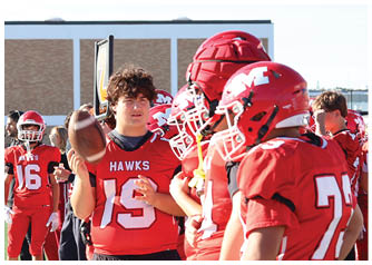A group of people wearing red football uniforms with helmets stand on the sidelines of a football field, with one person holding a football and wearing a jersey labeled “Hawks." Description generated by AI
