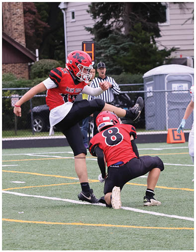A person in a red and black football uniform is kicking a football held by another person kneeling on a turf field with a referee in the background. Description generated by AI