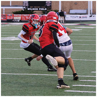 Three people wearing football helmets and uniforms are engaged in a play on a football field with a scoreboard showing one team leading. Description generated by AI