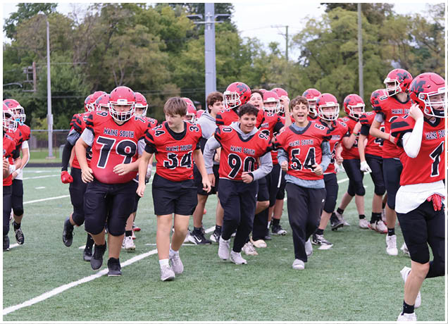A group of people wearing red and black football uniforms with helmets are walking together on a green field. Description generated by AI