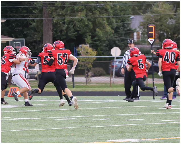 Several people in red and white football uniforms are actively playing on a green field with a referee and a down marker showing the number three in the background. Description generated by AI