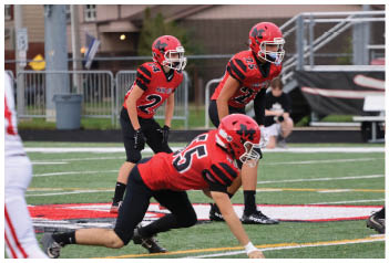 Four people in red and black football uniforms are positioned on a football field facing a person in a white and red uniform, appearing ready for a play. Description generated by AI