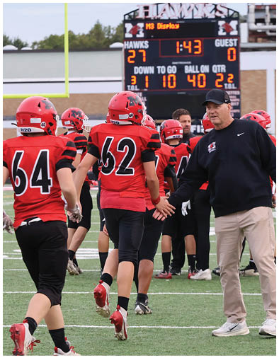 People in red and black football uniforms are walking on a field toward a scoreboard showing the score and time remaining. Description generated by AI