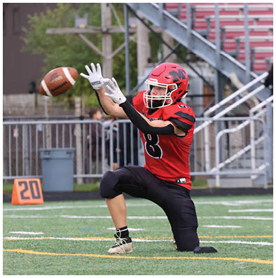 A person wearing a red and black football uniform is kneeling on one knee with arms extended, preparing to catch a football on a field near a 20-yard marker. Description generated by AI