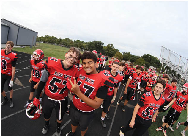 A group of people wearing red and black football uniforms are standing and smiling on a track field near bleachers and a building. Description generated by AI