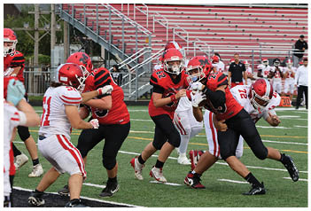 Players in red and white football uniforms are actively engaged in a tackle on a green field with bleachers and other players in the background. Description generated by AI