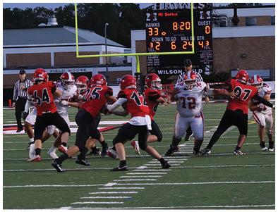 A group of people wearing football uniforms are playing on a field with a scoreboard and goalposts visible in the background during a game. Description generated by AI