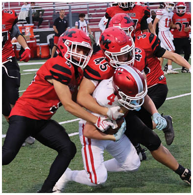 Three football players in red and black uniforms tackle a player in a white and red uniform on a green field during a game. Description generated by AI
