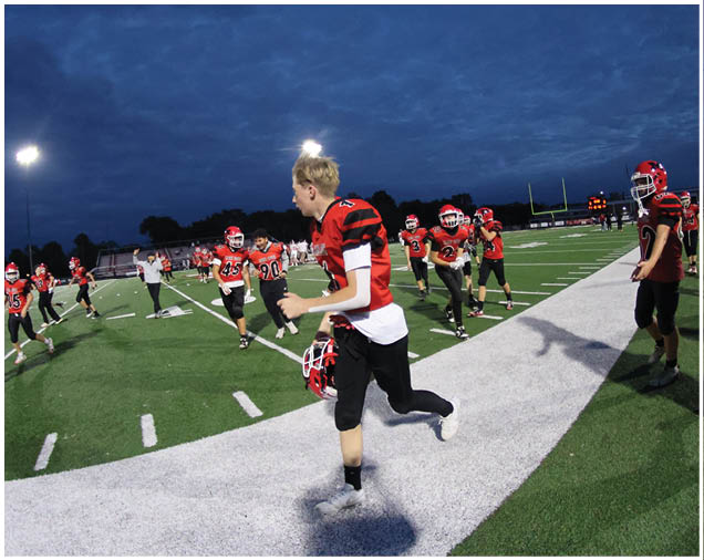 People wearing red and black football uniforms are on a field at dusk, with some running and others standing near the sideline. Description generated by AI