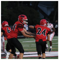 Two people in red football uniforms with helmets are engaged in a play on a football field while a person in a white uniform stands in the background. Description generated by AI