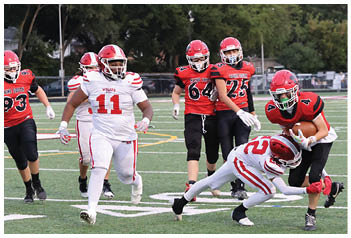 Two people in red football uniforms are on a field with one holding a football while being tackled by a person in a white uniform, with two others in red uniforms watching nearby. Description generated by AI