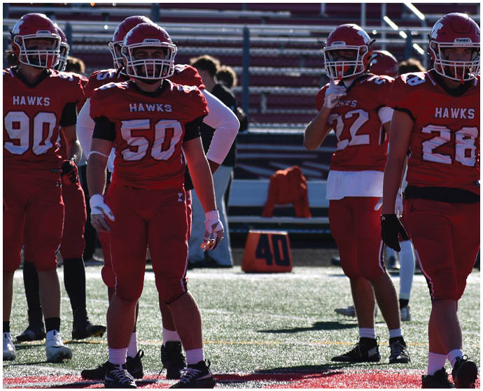 Four people in red football uniforms with helmets stand on a field near a yard marker and bleachers. Description generated by AI