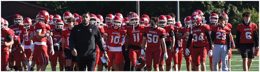 A group of people wearing red football uniforms and helmets are gathered on a field with one person in black clothing standing among them. Description generated by AI