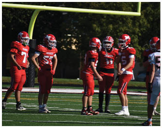 Two people wearing red football uniforms stand on a green field with white and yellow lines. Description generated by AI